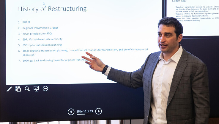 Associate Professor Joshua Macey ’17 stands in a classroom in front of a large projector screen. A list on the screen has the heading "History of Restructuring."