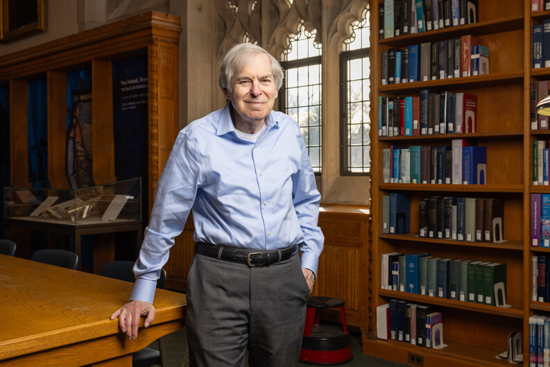 Fred Shapiro stands alongside a table and in front of a wall of bookshelves a multipane arched window framed with stone or concrete ornament