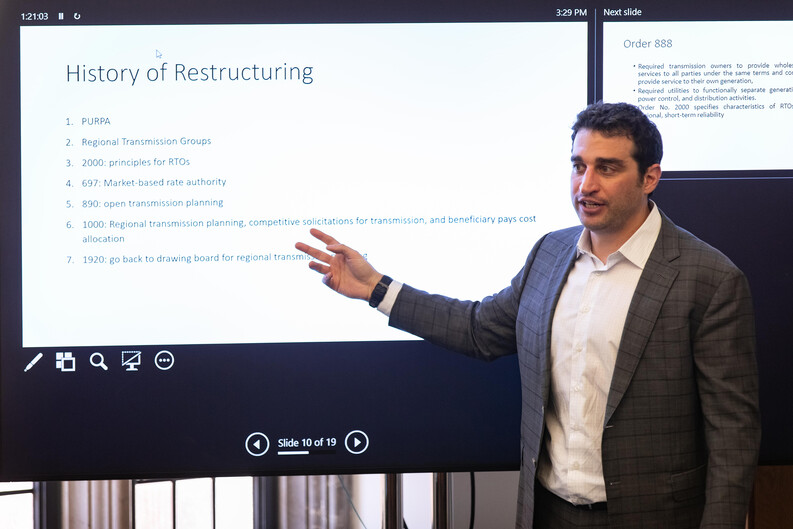 Associate Professor Joshua Macey ’17 stands in a classroom in front of a large projector screen. A list on the screen has the heading "History of Restructuring."