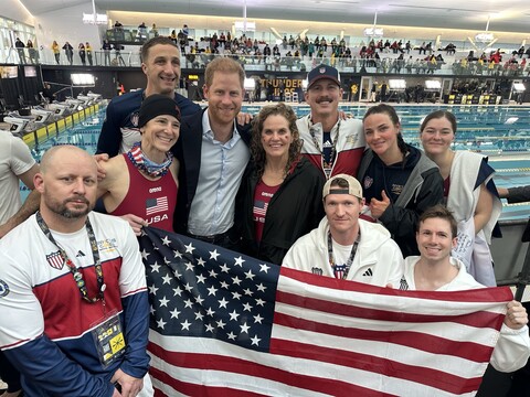 Swim team with Price Harry A group of people holding a U.S. flag pose for a photo by a swimming pool