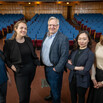 Five people pose for a group photo standing in front of a rows of auditorium seating
